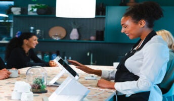 Woman smiling at POS machine with customers in background. Source: Tech.co