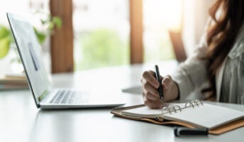 A woman sits at her desk, watching an online course and taking notes.