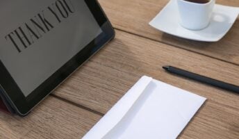 An ipad with the phrase thank you on it, next to a cup of coffee, pen, and envelope.